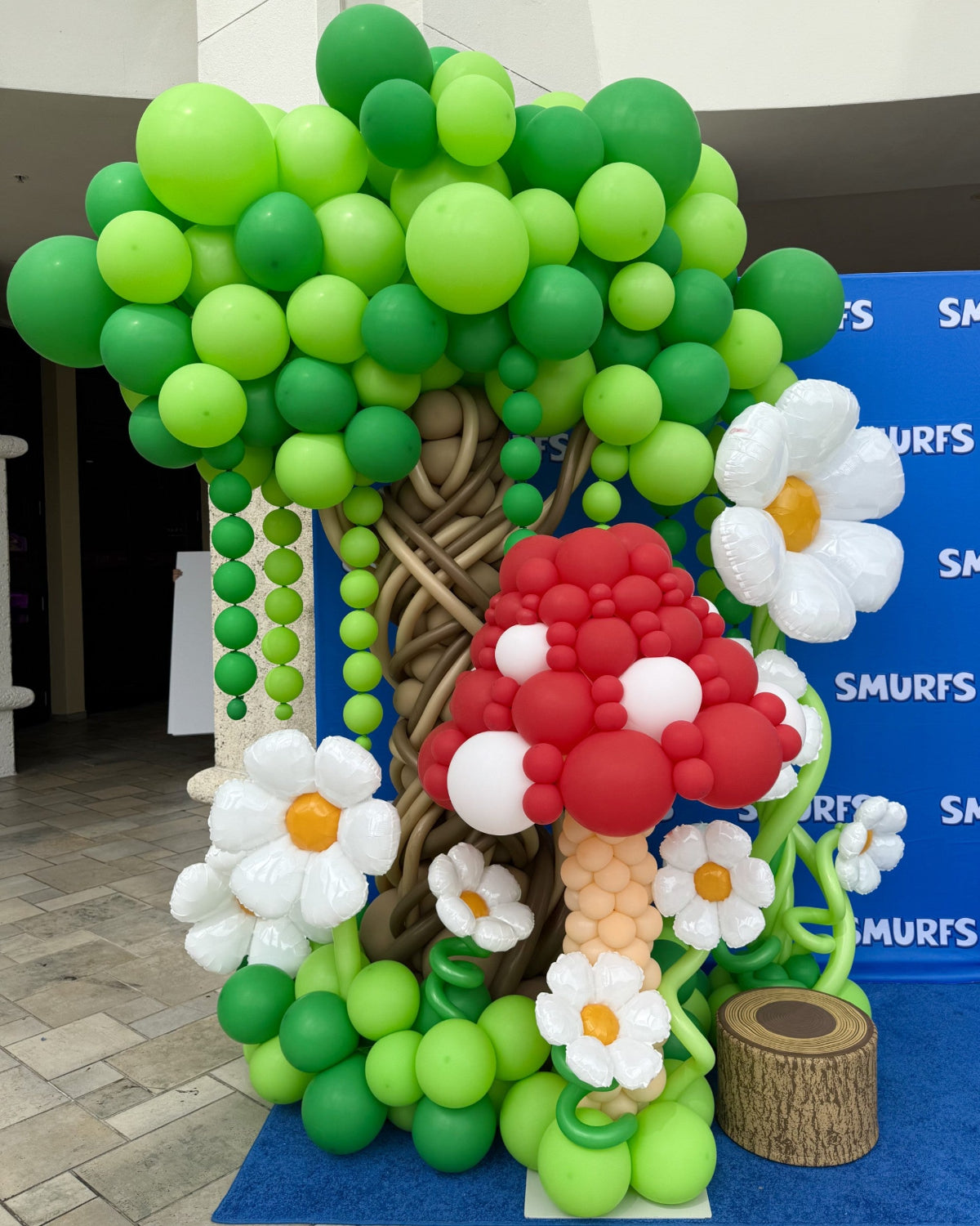 Whimsical balloon installation featuring a large green balloon tree with twisted brown trunk, a red-and-white balloon mushroom, and scattered balloon daisies with yellow centers. The base is filled with green balloon foliage and a wood stump prop, all set against a Smurfs-themed backdrop. Created as part of The Creative Heart Studio’s Mushroom Balloon Class & Recipes, this setup is perfect for storybook parties and immersive event decor.