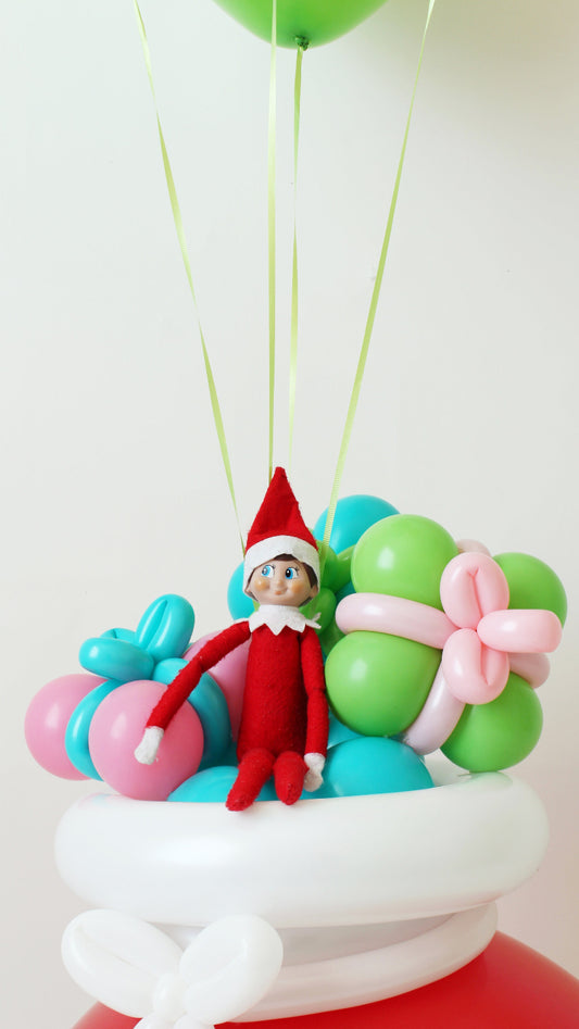 Close-up of Elf on the Shelf sitting atop a white balloon sack lid, surrounded by pastel balloon presents in pink, green, teal, and blue. Part of Lilly Jimenez’s Elf Arrival Santa Sack tutorial, this creative balloon design makes the perfect whimsical touch for Elf arrival displays, holiday decor, or Christmas morning surprises.