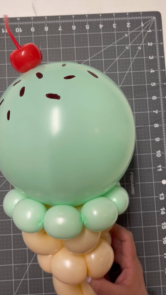 Balloon sculpture of a mint green ice cream cone with a red cherry on top, chocolate sprinkle details, and a beige cone base, placed on a cutting mat. Perfect for summer events, party centerpieces, dessert-themed décor, and balloon artist creations.