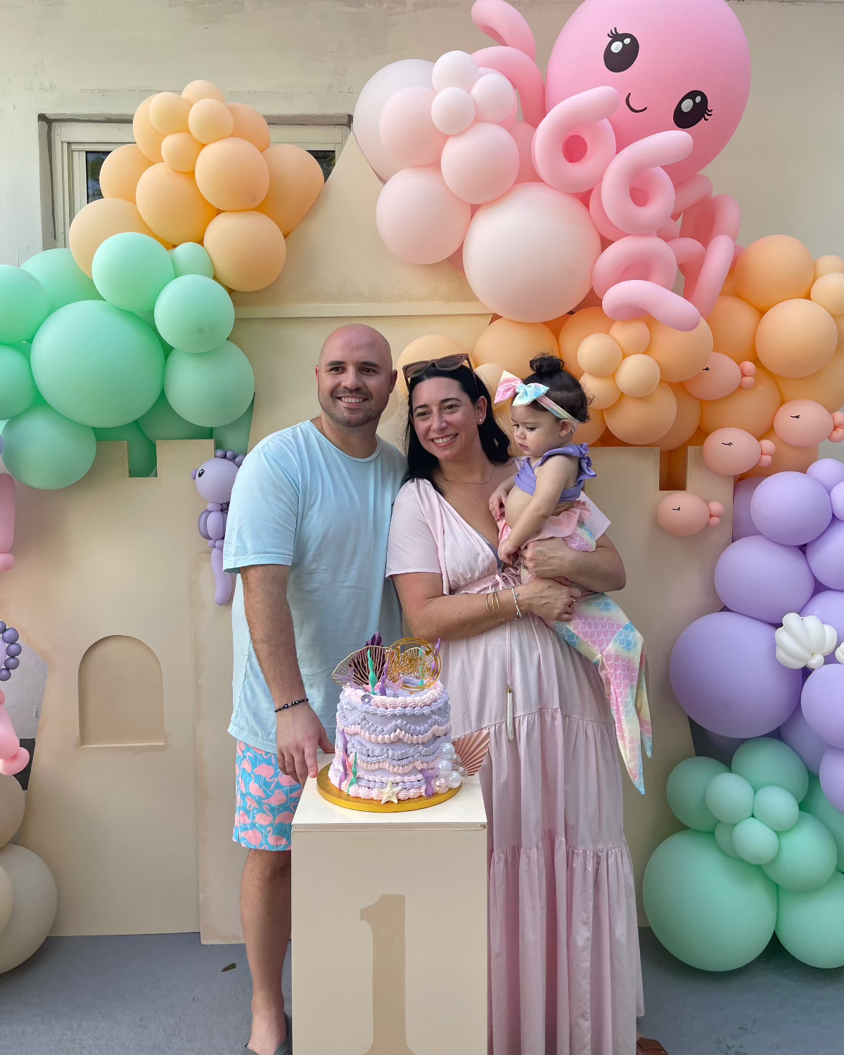 Mom and dad celebrate their daughter’s first birthday with a pastel balloon-decorated foam board Sand Castle Backdrop. The scene features a large pink balloon octopus, ocean-themed garlands, and a birthday cake on a “1” pedestal. This whimsical setup is part of the Sand Castle Backdrop Class by The Creative Heart Studio—perfect for beach, mermaid, or under-the-sea themed parties.
