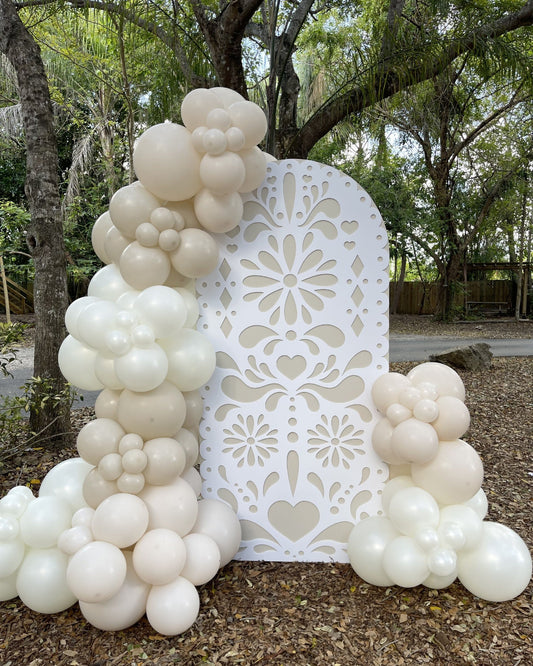 Large white papel picado-style foam board backdrop decorated with cream and ivory balloon clusters. Displayed outdoors among trees, this elegant party setup is ideal for weddings, baby showers, birthdays, or Día de Muertos events. Design made using a laser cut or CNC machine with files available in SVG, DXF, and printable formats.