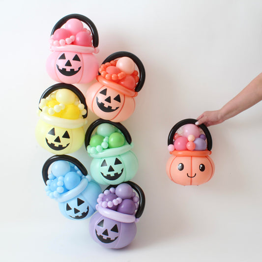 A set of pastel-colored balloon jack-o'-lantern candy buckets, each filled with balloon "candies" and topped with a black balloon handle. The colors range from pink, orange, yellow, green, blue, and purple, creating a fun and modern Halloween aesthetic. A hand holds a cute smiling pumpkin bucket, adding a playful touch. Designed by Lilly Jimenez of The Creative Heart Studio, these balloon creations are perfect for Halloween parties, trick-or-treat events, and festive displays.