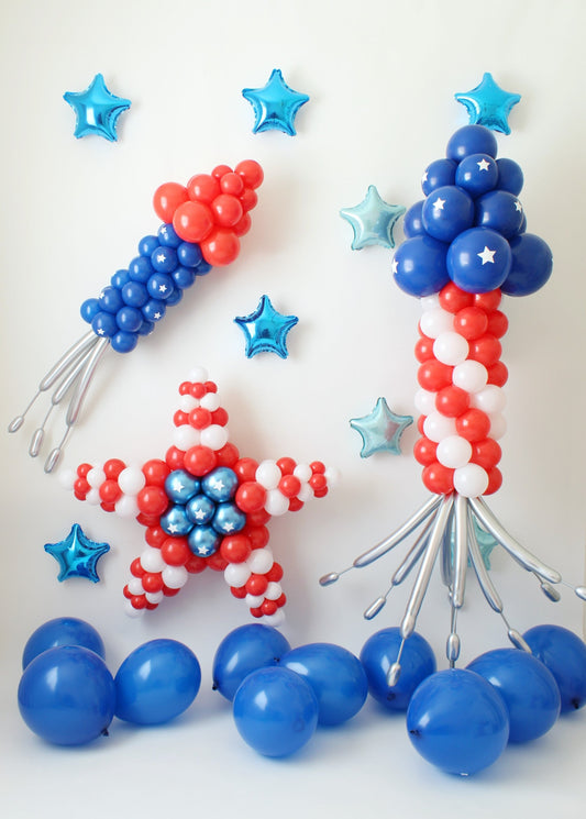 A festive red, white, and blue balloon setup featuring a balloon star, a small bottle rocket, and a large bottle rocket column, accented with metallic blue star balloons and scattered blue balloons on the floor. Designed by Lilly Jimenez of The Creative Heart Studio, this patriotic display is perfect for Fourth of July celebrations, parades, and patriotic-themed events. 🎆🎈