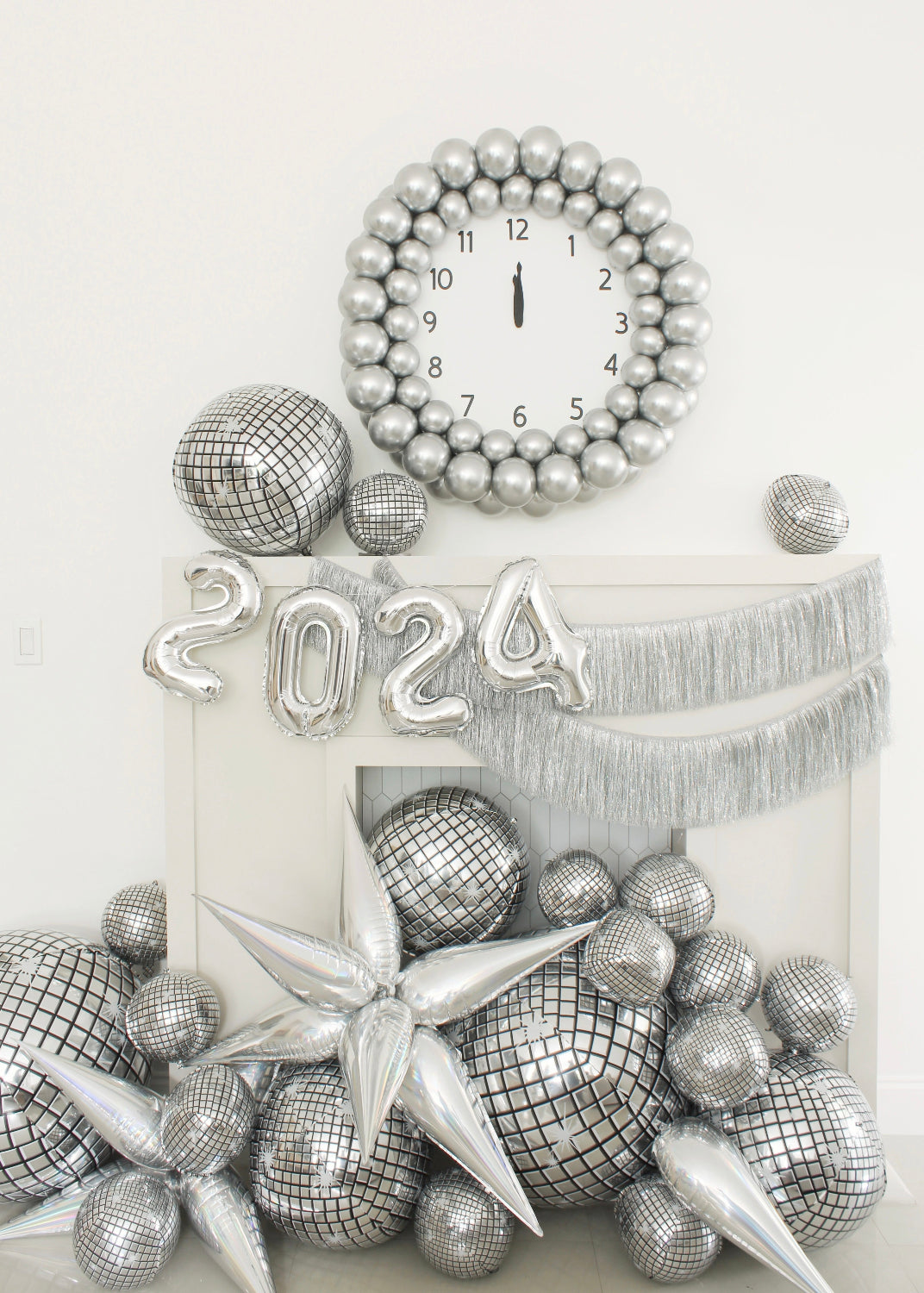 Stylish New Year’s Eve 2024 balloon decor featuring a large silver balloon clock striking midnight, reflective disco balloons, silver balloon numbers “2024,” metallic starbursts, and shimmering fringe garland. This festive setup by Lilly Jimenez is perfect for NYE parties, balloon artists, or anyone looking to create stunning photo ops with creative balloon installations.