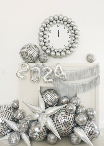 Stylish New Year’s Eve 2024 balloon decor featuring a large silver balloon clock striking midnight, reflective disco balloons, silver balloon numbers “2024,” metallic starbursts, and shimmering fringe garland. This festive setup by Lilly Jimenez is perfect for NYE parties, balloon artists, or anyone looking to create stunning photo ops with creative balloon installations.