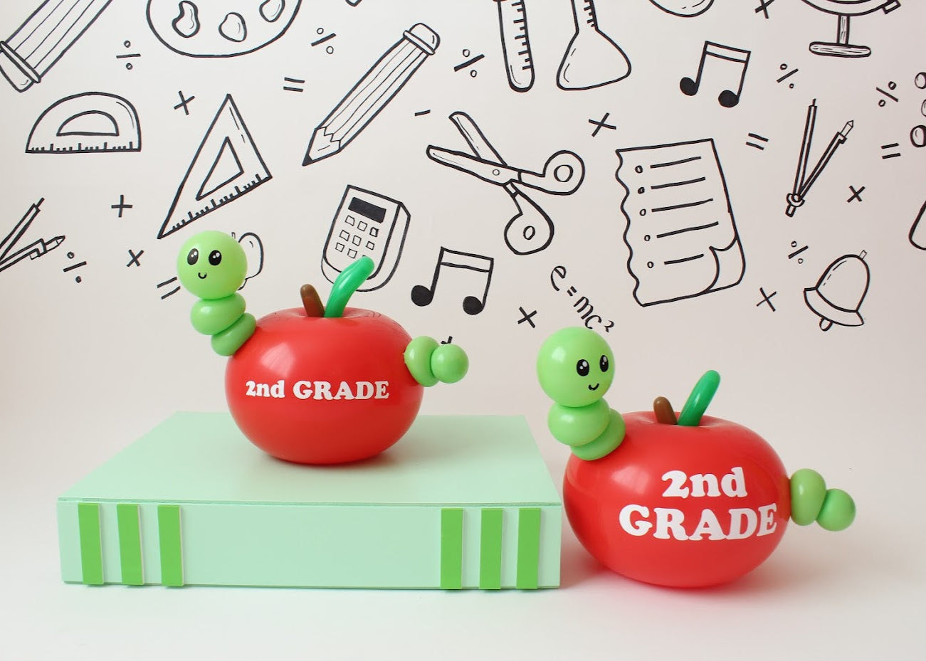 Two red balloon apples labeled "2nd GRADE," each featuring an adorable green balloon worm, sit on a green book-shaped prop against a white backdrop decorated with hand-drawn school-themed doodles. The playful design makes for a fun and creative back-to-school decoration.