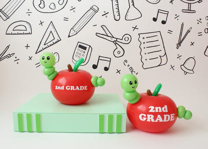 Two red balloon apples labeled "2nd GRADE," each featuring an adorable green balloon worm, sit on a green book-shaped prop against a white backdrop decorated with hand-drawn school-themed doodles. The playful design makes for a fun and creative back-to-school decoration.