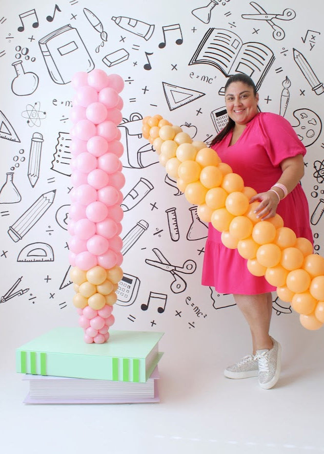 Lilly Jimenez poses with two creative back-to-school balloon columns against a white backdrop decorated with hand-drawn school-themed doodles. One column, made of pink and beige balloons, stands atop a stack of oversized book props, while the other, designed with orange balloons, is shaped like a giant colored pencil. She wears a bright pink dress and smiles while holding the orange pencil balloon column.