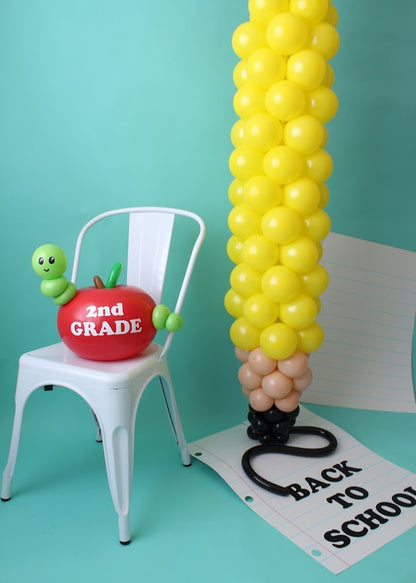 A festive back-to-school balloon display featuring a tall pencil-shaped balloon column with a yellow body, brown and black tip, and a pink eraser top. Next to it, a white metal chair holds a red balloon apple labeled "2nd Grade," complete with a green balloon worm. A large sheet of balloon-crafted notebook paper on the floor displays "Back to School" text, all set against a teal background.