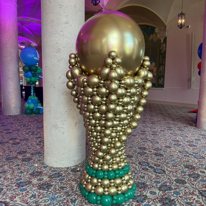 Giant gold balloon soccer trophy sculpture with green balloon base displayed indoors at a FIFA World Cup event venue.