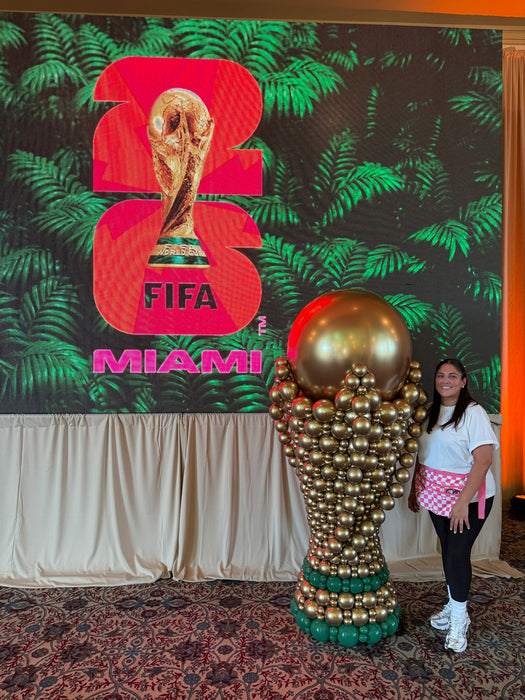 Lilly Jimenez standing next to a giant gold balloon soccer trophy sculpture in front of a FIFA World Cup 2026 Miami event display.