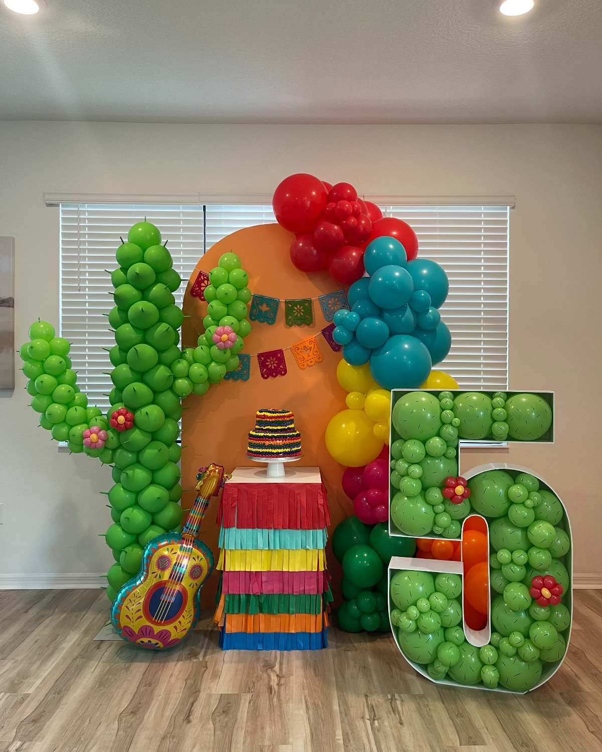 Colorful fiesta-themed balloon setup featuring a tall green balloon cactus column, a number 5 balloon marquee filled with mini green balloons and flowers, vibrant balloon garland in red, yellow, and teal, and a rainbow fringe-covered table with a layered cake topper—perfect for Cinco de Mayo or a fiesta birthday party backdrop.