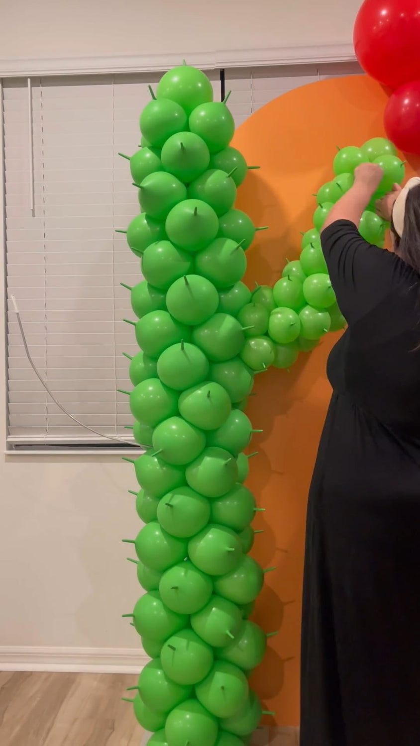 Balloon artist assembling a large green balloon cactus column using round balloons and 260Qs for spikes. The cactus structure is part of a vibrant fiesta backdrop with an orange panel and red balloon cluster—perfect for Cinco de Mayo, southwestern, or desert-themed parties and events.