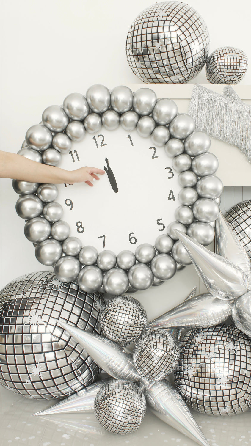 Close-up of a New Year’s Eve balloon clock made with metallic silver balloons and a printed clock face showing midnight. A hand is seen adjusting the minute hand. Surrounded by reflective disco balloon decor and silver starburst accents, this photo captures a key moment from Lilly Jimenez’s New Year’s Eve Clock Tutorial—perfect for balloon artists planning eye-catching NYE installations.