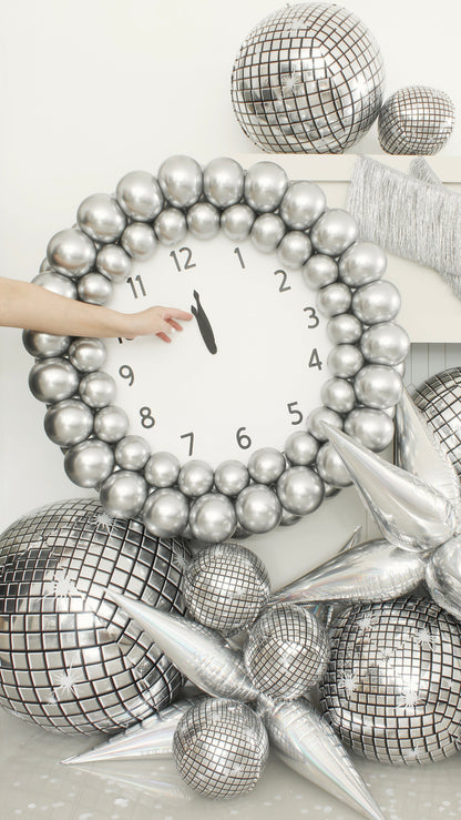Close-up of a New Year’s Eve balloon clock made with metallic silver balloons and a printed clock face showing midnight. A hand is seen adjusting the minute hand. Surrounded by reflective disco balloon decor and silver starburst accents, this photo captures a key moment from Lilly Jimenez’s New Year’s Eve Clock Tutorial—perfect for balloon artists planning eye-catching NYE installations.