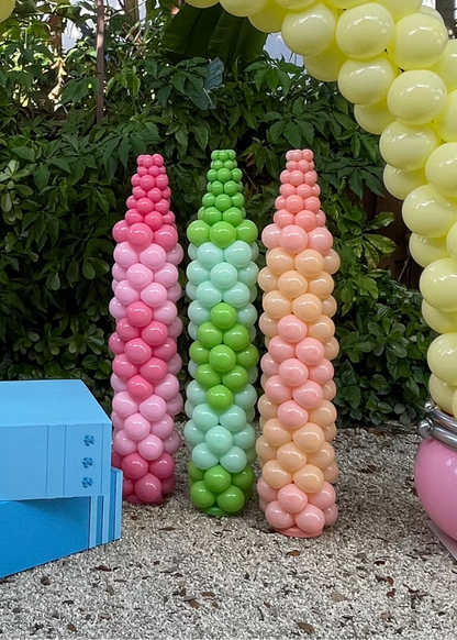 Outdoor display of three colorful Balloon Crayon Columns in pink, green, and orange, part of the Back to School Column Class. These school-themed balloon sculptures are ideal for first day of school events, classroom decor, and teacher appreciation setups. The class includes video tutorials and printable balloon recipes for balloon artists and event stylists.