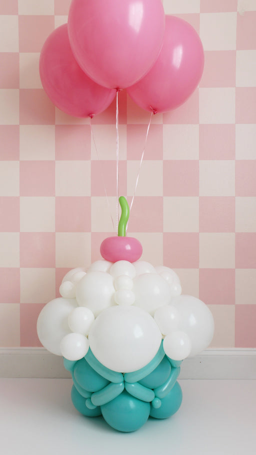 A large balloon cupcake sculpture featuring a teal base, white frosting made of balloons, and a pink balloon cherry with green stem, topped with three floating pink balloons. This whimsical balloon design is perfect for birthdays, candyland parties, dessert table decor, and themed balloon displays.