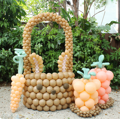 Outdoor setup featuring a giant balloon Easter bunny basket with floppy ears, bunny face details, and large sprouting balloon carrots in pastel orange and pink. Perfect for Easter photo backdrops and festive spring balloon displays.
