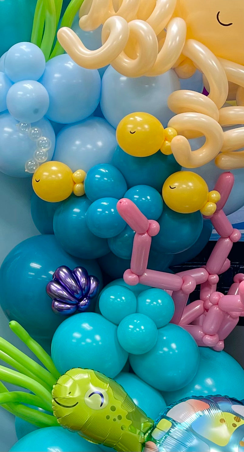 Balloon sculpture featuring small yellow balloon fish, a metallic purple seashell, seaweed, and ocean-inspired balloon clusters. Part of the Under the Sea Balloon Class, this vibrant display adds a playful touch to any underwater-themed party or event décor.