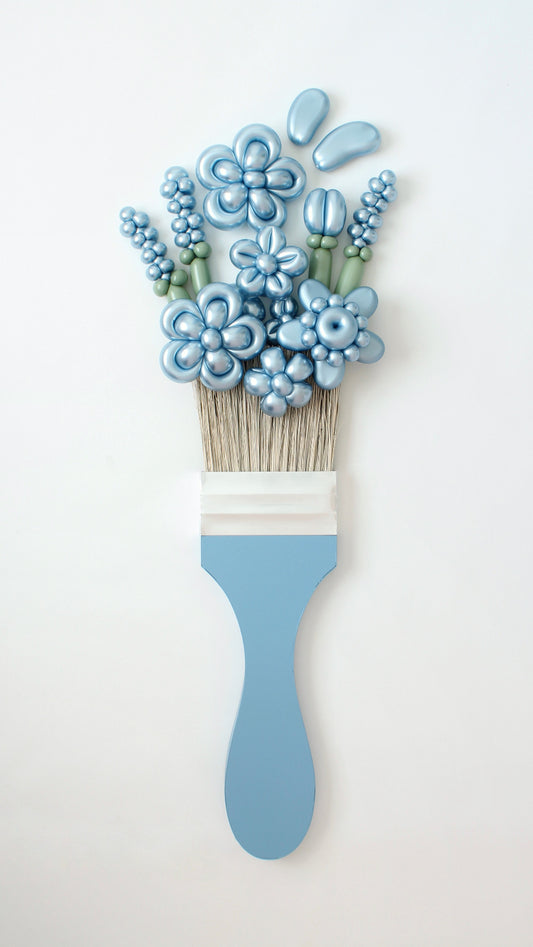An oversized paintbrush prop with bristles that appear to be "painting" a stunning arrangement of metallic blue balloon flowers and greenery. The unique design creates an artistic illusion, blending balloon artistry with a floral aesthetic. Designed by Lilly Jimenez of The Creative Heart Studio, this piece is perfect for art-themed events, creative decor, and photoshoots.