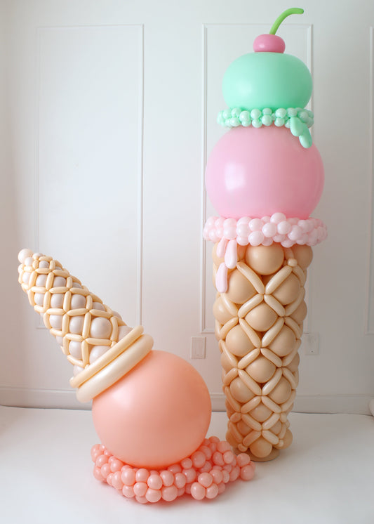 Two large balloon ice cream cone sculptures against a white-paneled wall. One design features a towering double-scoop cone with pastel pink and green scoops, a cherry on top, and a realistic balloon waffle cone base. The second design is a playful upside-down ice cream cone with a peach-colored scoop, a detailed waffle-textured cone, and a pink balloon cherry, appearing to have spilled onto a cluster of smaller balloons.