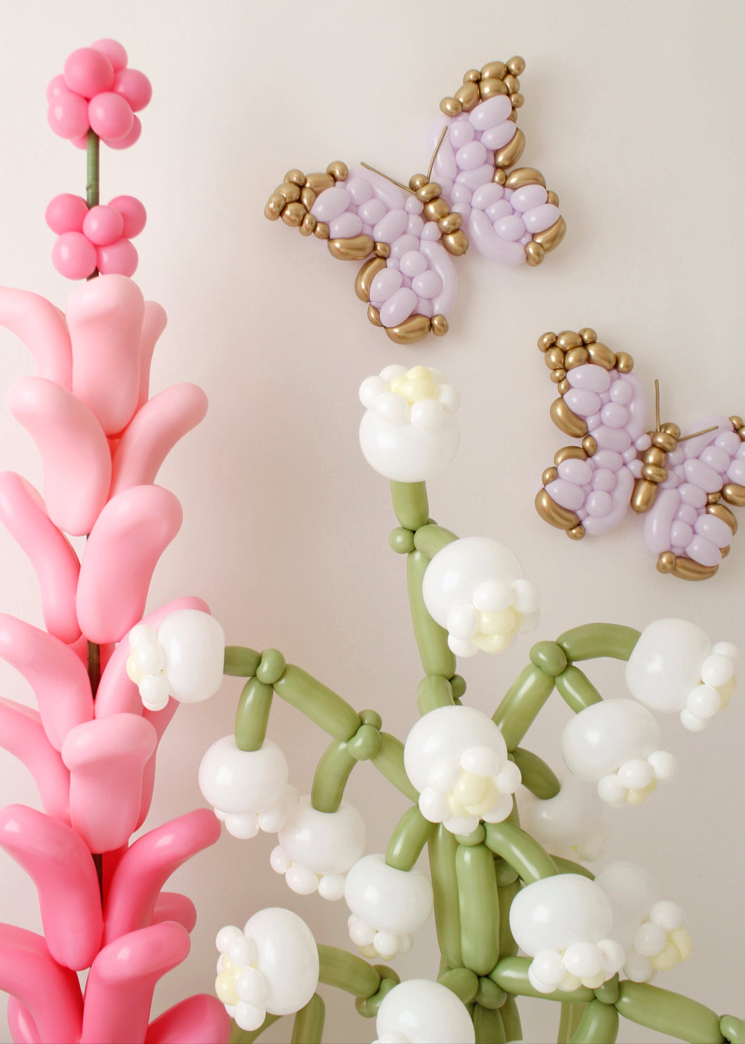 Elegant balloon lilies of the valley with delicate white blooms, pink balloon lavender, and handcrafted balloon butterflies in soft lavender with gold accents. Set against a neutral backdrop, this whimsical arrangement adds a magical, enchanted garden feel, perfect for event decor, fairy tale themes, and balloon artistry inspiration.