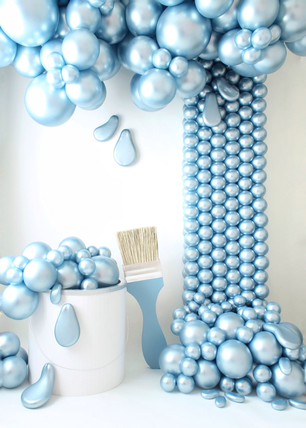 A creative balloon installation featuring an oversized paint bucket prop overflowing with metallic blue balloon "paint", cascading down in a dramatic spill effect. A matching oversized paintbrush rests beside the bucket, completing the artistic setup. Designed by Lilly Jimenez of The Creative Heart Studio, this setup is perfect for art-themed events, photoshoot backdrops, and creative decor displays.