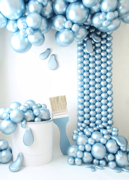 A creative balloon installation featuring an oversized paint bucket prop overflowing with metallic blue balloon "paint", cascading down in a dramatic spill effect. A matching oversized paintbrush rests beside the bucket, completing the artistic setup. Designed by Lilly Jimenez of The Creative Heart Studio, this setup is perfect for art-themed events, photoshoot backdrops, and creative decor displays.