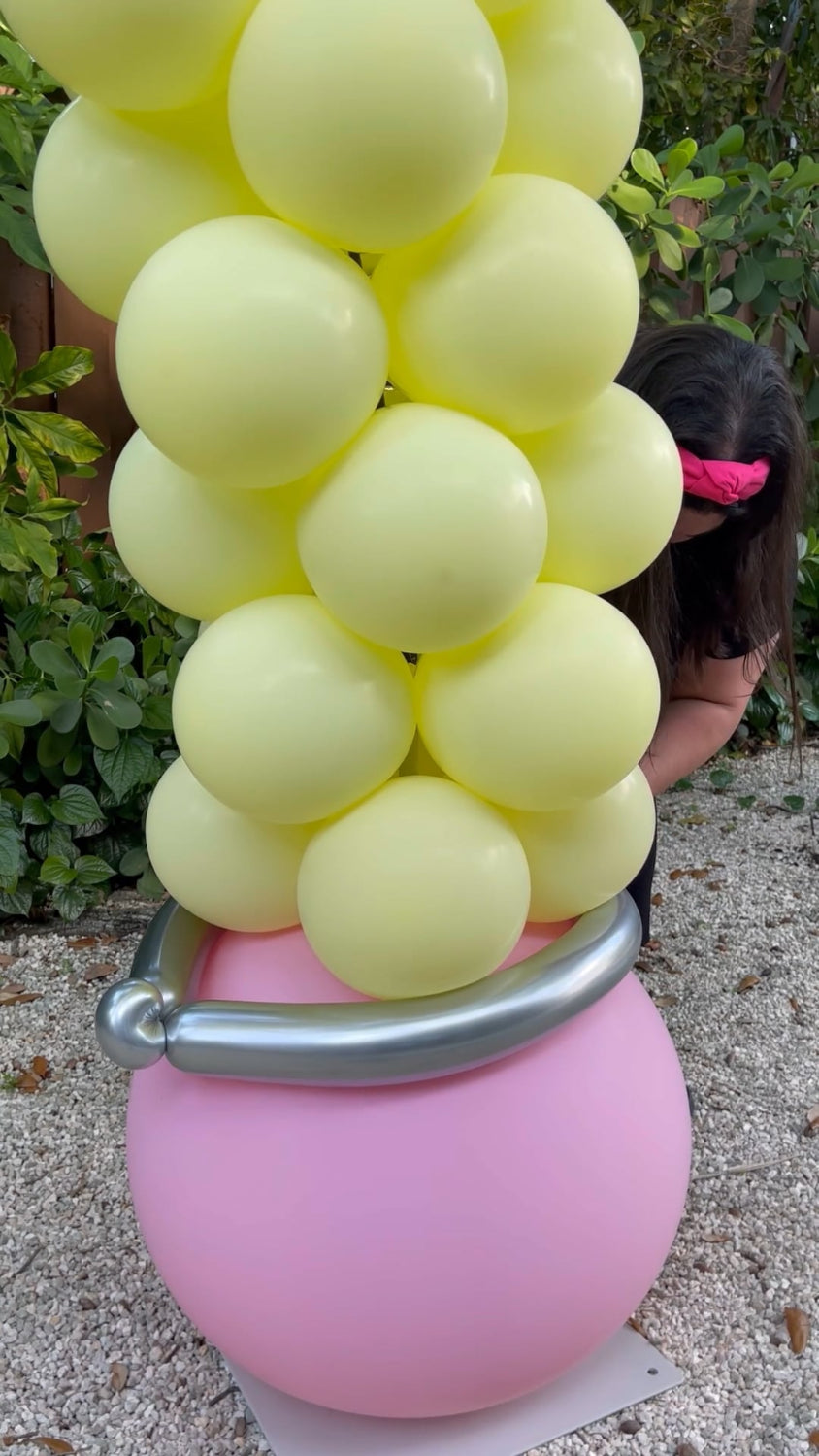 Close-up of the eraser base of a pencil balloon arch, featuring pastel yellow balloons stacked above a large pink balloon with a metallic silver band to resemble a pencil eraser. A woman is partially visible adjusting the base. Ideal for back-to-school, teacher appreciation, and classroom decoration tutorials.