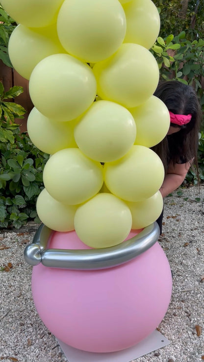 Close-up of the eraser base of a pencil balloon arch, featuring pastel yellow balloons stacked above a large pink balloon with a metallic silver band to resemble a pencil eraser. A woman is partially visible adjusting the base. Ideal for back-to-school, teacher appreciation, and classroom decoration tutorials.