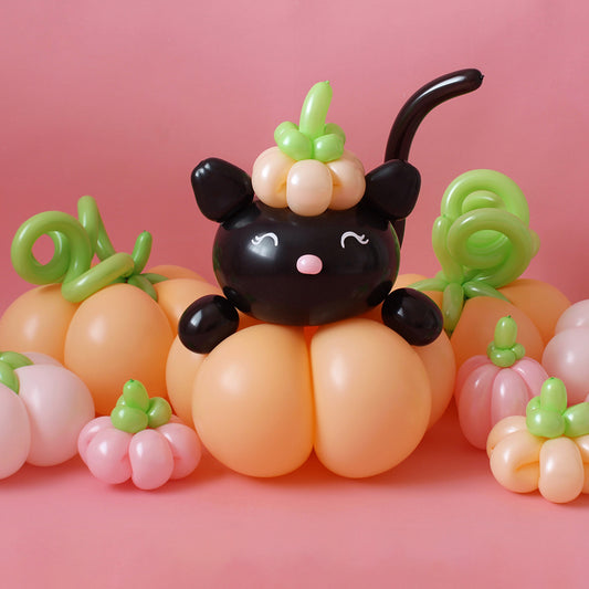 A charming balloon display featuring an adorable black cat sitting on a stack of pastel and orange balloon pumpkins, complete with curly green vines. The cat wears a mini pumpkin on its head, adding a playful and festive touch. This whimsical design is perfect for Halloween, fall festivals, and seasonal party decorations. Learn to create this fun and eye-catching setup in the Balloon Pumpkin & Cat Tutorial!