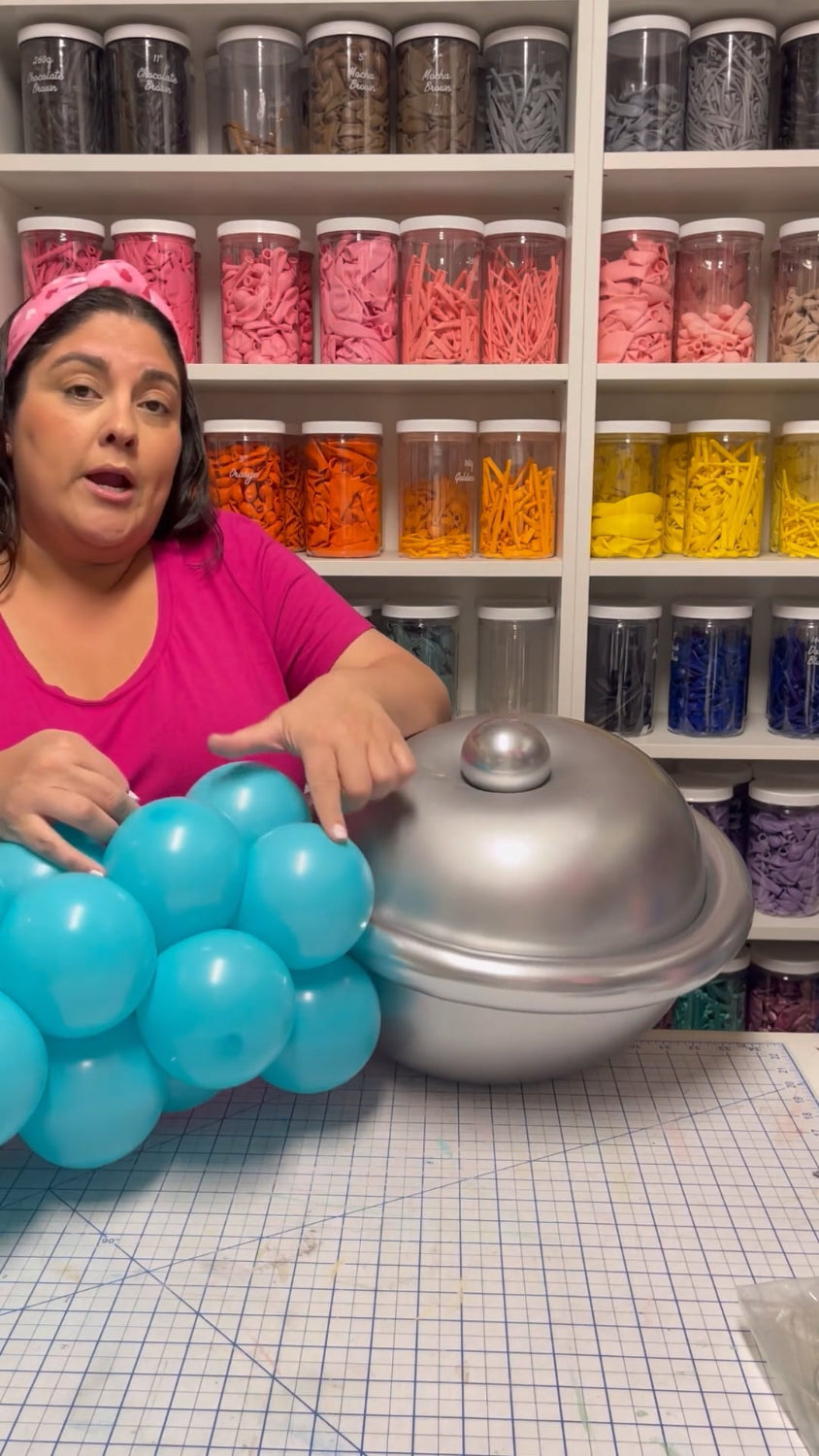 Lilly demonstrates how to attach the metallic balloon chest piece for a stethoscope sculpture in her tutorial, surrounded by organized balloon supplies in her craft room.