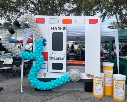 A large balloon stethoscope sculpture is displayed on an ambulance-themed photo booth backdrop at an outdoor event, with oversized prescription bottles and themed signage for a medical celebration.