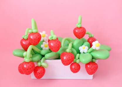 A white basket filled with balloon strawberries, green balloon leaves, and small balloon daisies on a pink background, creating a cheerful garden-inspired balloon arrangement.