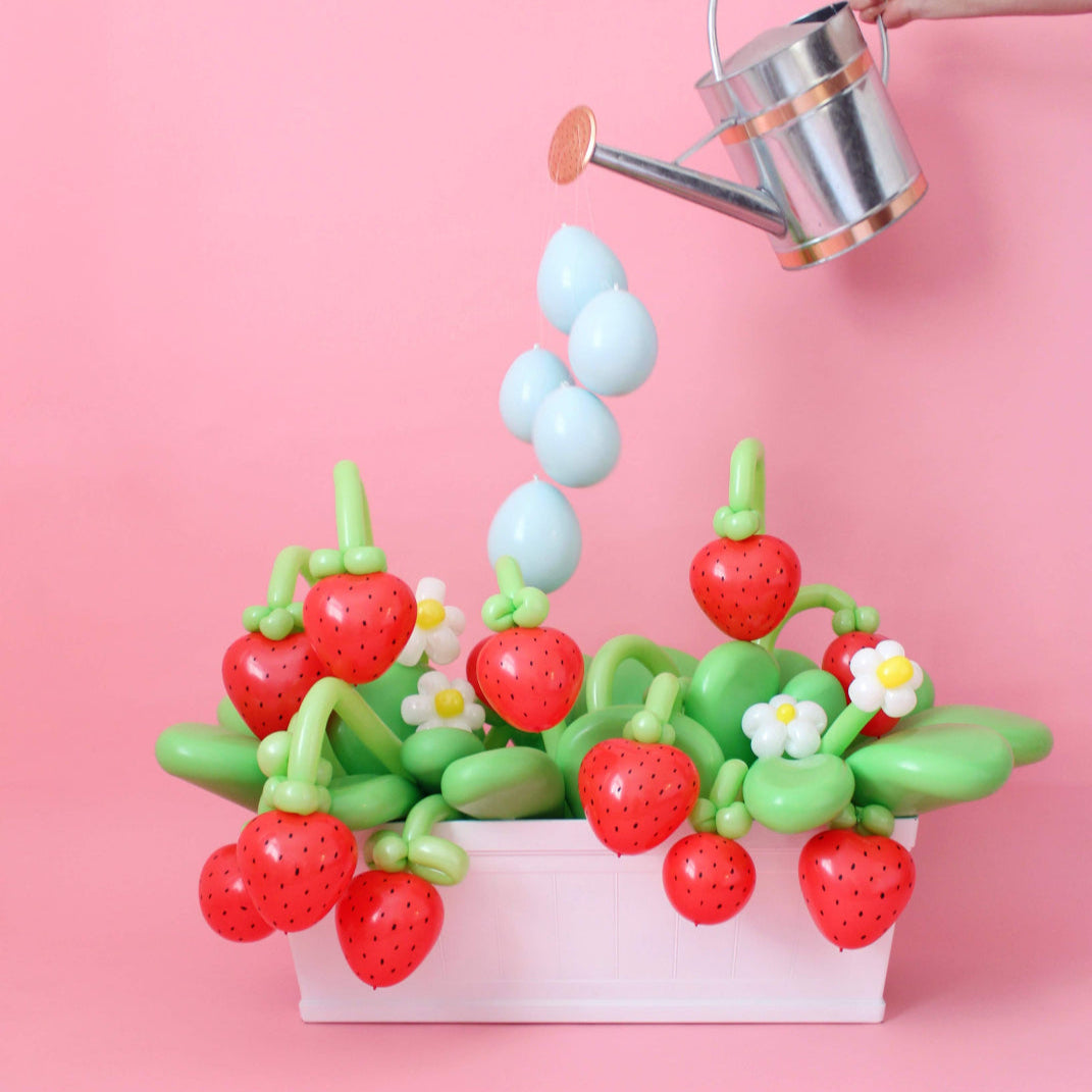 A playful balloon strawberry garden display featuring red balloon strawberries, green balloon leaves, white and yellow daisies, and a hand holding a watering can with blue balloons resembling water droplets, all set against a pink backdrop.