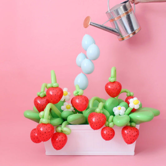 A playful balloon strawberry garden display featuring red balloon strawberries, green balloon leaves, white and yellow daisies, and a hand holding a watering can with blue balloons resembling water droplets, all set against a pink backdrop.
