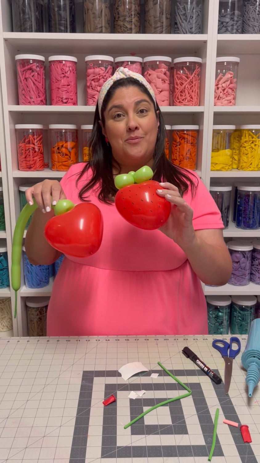 Lilly Jimenez in a pink dress demonstrates how to make balloon strawberries, holding two red balloon strawberries with green balloon tops in a colorful craft room.