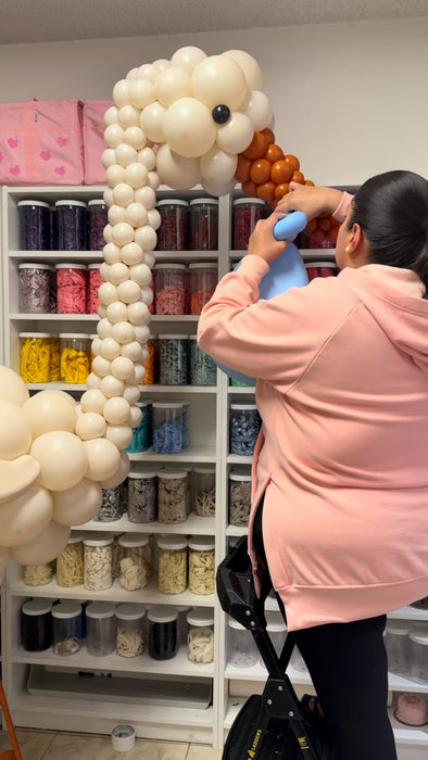Behind the scenes photo of a balloon artist assembling a large stork balloon sculpture in a studio, showing the construction process for a professional welcome baby balloon design.