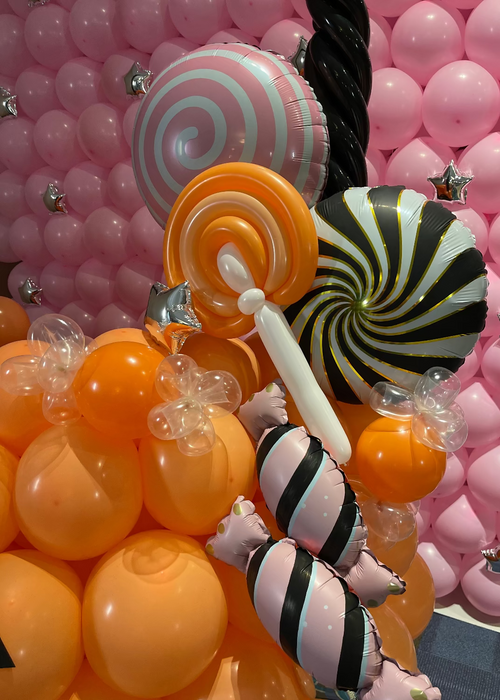 Close-up of balloon candy decorations on a balloon pumpkin treat bucket. The design features orange balloons forming the pumpkin, with oversized spiral lollipop balloons in pink, orange, and black, plus striped candy balloons. A twisted black balloon handle and a pink balloon wall backdrop complete this festive Halloween balloon decor perfect for parties, schools, and photo backdrops.