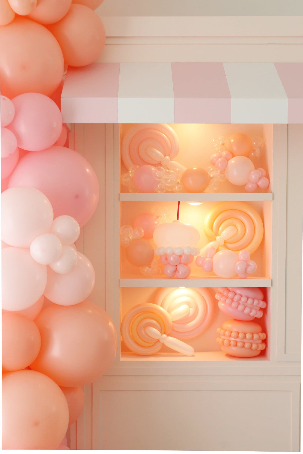 Close-up of a foamboard storefront display featuring balloon candy props, including lollipops, macarons, and a cupcake, all styled in pastel pinks and peaches. A pink-and-white striped awning tops the structure, with shelves lit from behind for an eye-catching party prop. Part of the Foamboard Storefront Class from The Creative Heart Studio.