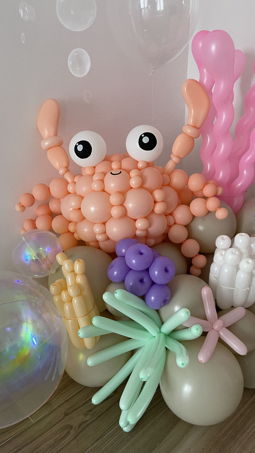 Balloon crab sculpture created using peach and orange balloons, surrounded by sea-inspired elements like mint sea plants, bubble balloons, coral, and underwater textures. This setup is part of Lilly Jimenez’s Under the Sea Balloon Class and is ideal for beach parties, mermaid themes, and aquatic decor. The tutorial teaches decorators how to build this eye-catching ocean character step-by-step.