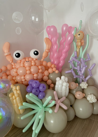 Ocean-inspired balloon display featuring a large peach balloon crab, coral, sea plants, seashells, and floating bubble balloons. Designed by Lilly Jimenez for the Under the Sea Balloon Class, this setup showcases various sculpted elements taught in the tutorial, including layered coral, underwater textures, and sea creatures—ideal for mermaid parties, ocean backdrops, and themed balloon installs.