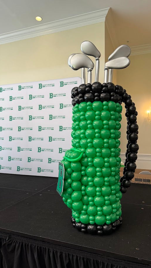 Large balloon golf bag structure made with green and black balloons, topped with silver balloon golf clubs and displayed on a stage at a branded event backdrop. Professional golf themed balloon decor used for tournaments, corporate events, and golf celebrations by The Creative Heart Studio.