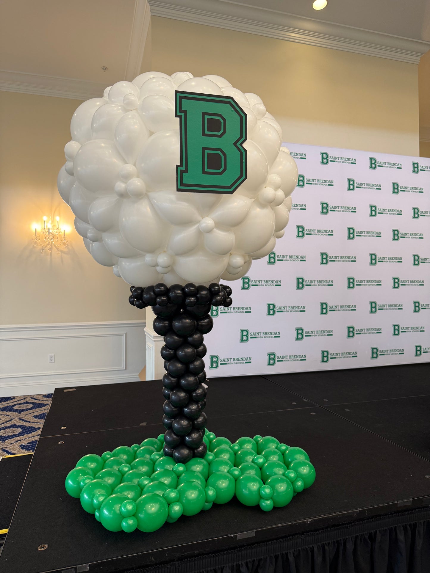 Balloon golf ball on a tee structure made with white, black, and green balloons, displayed on an indoor event stage with a branded backdrop. This professional golf themed balloon structure is designed for tournaments, corporate events, and stage decor, showcasing large scale balloon structure techniques by The Creative Heart Studio.