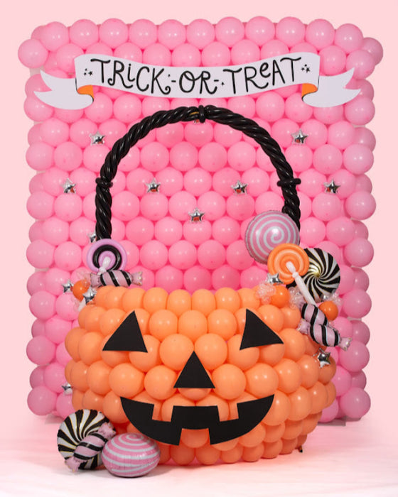 A balloon pumpkin bucket photo op featuring an oversized jack-o-lantern made of orange balloons with black face details, decorated with balloon candies and a black twisted handle. The backdrop is a wall of pink balloons with a white “Trick or Treat” banner, making this a playful and eye-catching Halloween balloon display for parties, schools, and festive events.