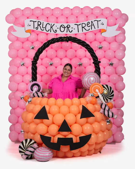 A large balloon pumpkin bucket photo op decorated with orange balloons, black jack-o-lantern face, and oversized balloon candies, set against a pink balloon wall with a “Trick or Treat” banner. Perfect Halloween balloon decor idea for parties, school events, and festive photo backdrops.