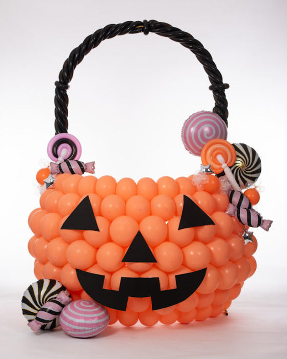 An oversized balloon pumpkin treat bucket made from orange balloons with a black jack-o-lantern face, twisted black balloon handle, and colorful balloon candies attached. This creative Halloween balloon design is perfect for festive displays, parties, school events, and seasonal photo backdrops.