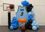 Basketball Jersey Backdrop featuring a custom light blue jersey with black trim, personalized with name and number, surrounded by blue and black balloon garlands. Includes balloon basketball hoops and Wilson basketball props for a complete sports photo setup. Perfect for pep rallies, senior nights, team banquets, and basketball-themed parties. Created using the Basketball Jersey Backdrop Template from The Creative Heart Studio.