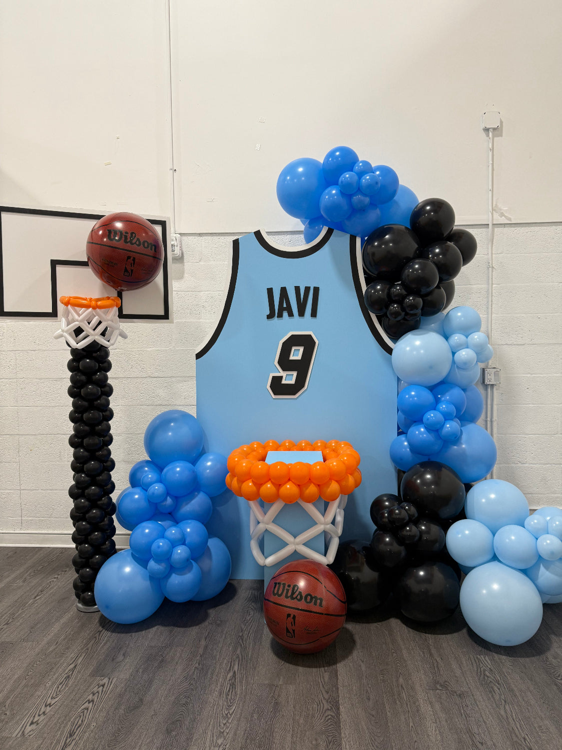 Basketball Jersey Backdrop featuring a custom light blue jersey with name and number, surrounded by black and blue balloon decor. Perfect for pep rallies, senior nights, and sports-themed celebrations. This design highlights school spirit and makes a bold photo backdrop for team events or athlete celebrations, created using the Basketball Jersey Backdrop Template from The Creative Heart Studio.