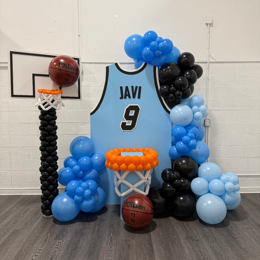 Basketball Jersey Backdrop featuring a custom light blue jersey with black trim, personalized name, and number centerpiece surrounded by blue and black balloon garlands. Complete with balloon basketball hoops and real Wilson basketballs, this setup is perfect for pep rallies, senior nights, team banquets, and basketball-themed events. Designed using the Basketball Jersey Backdrop Template from The Creative Heart Studio.
